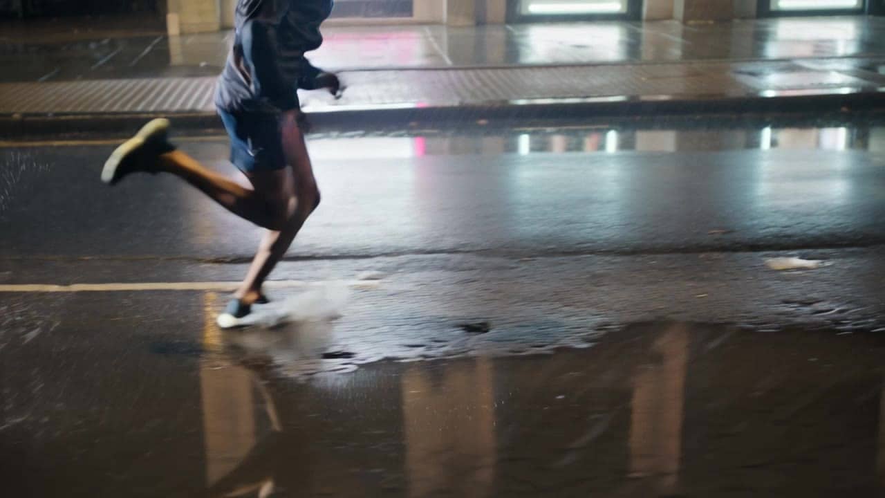 Kling 3 Standard AI video example: Wide 16:9 cinematic action shot, a runner sprints through a rainy city street at night, water splashes...
