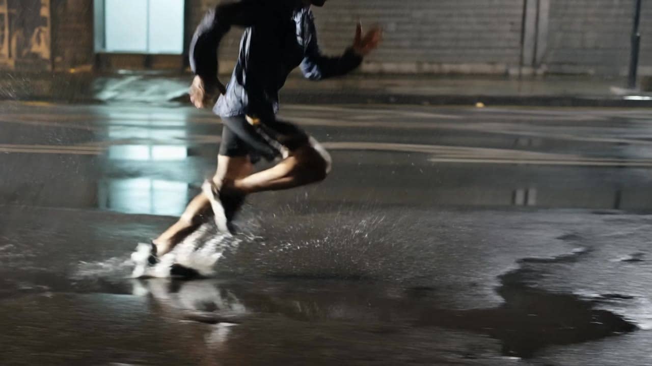 Ejemplo de video IA de Kling 3 Pro: Wide 16:9 cinematic action shot, a runner sprints through a rainy city street at night, water splashe...
