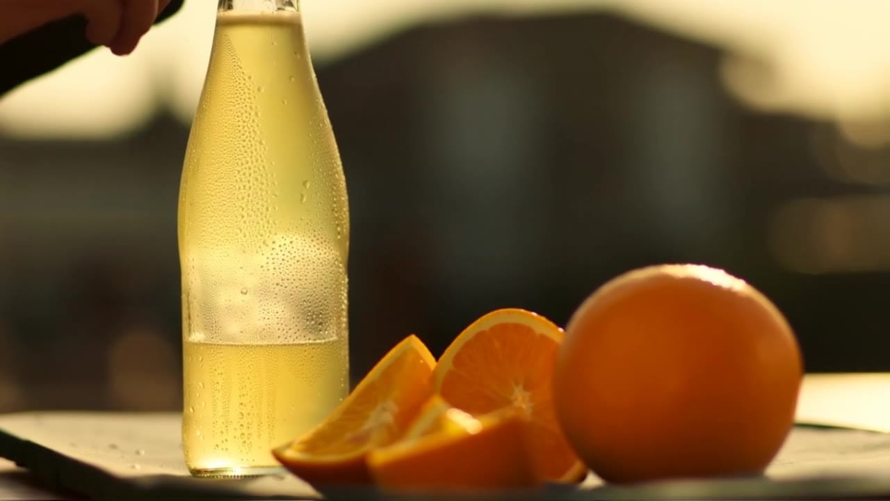 Ejemplo de video IA de Luma Ray 2: Scene (plain language): A glass bottle of sparkling citrus drink sits on a sunlit outdoor café table i...