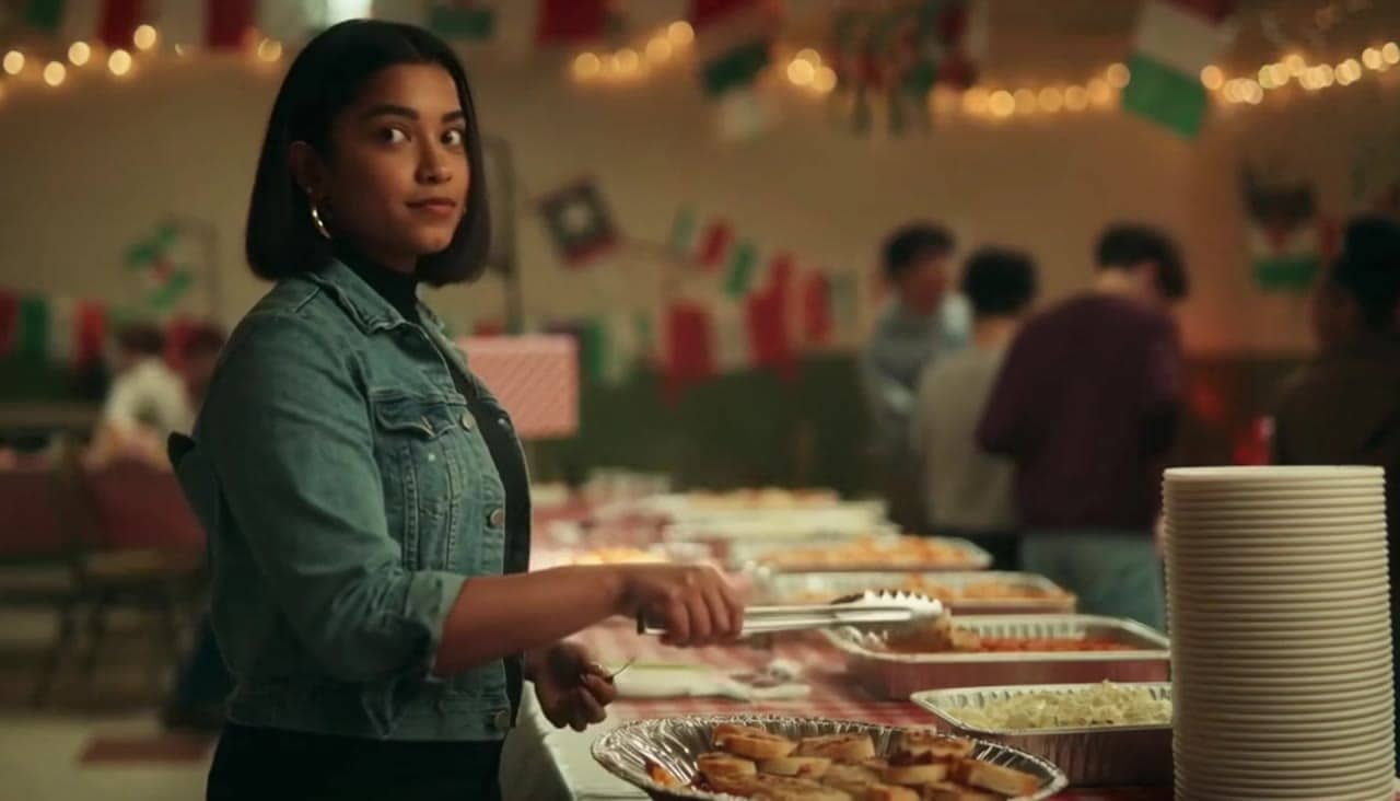 Ejemplo de video IA de OpenAI Sora 2 Pro: Bella sits at a long table draped in a red-and-white checkered tablecloth, a paper plate of hal...